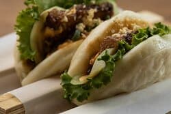 Hoisin Pork Belly Bao filled with hoisin glazed pork belly, cucumber, peanut dressing, chicharron, and lettuce.