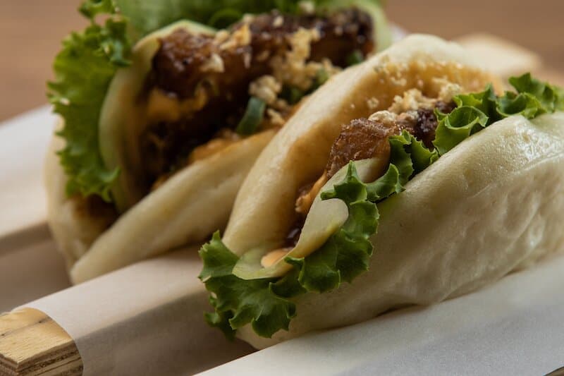 Hoisin Pork Belly Bao filled with hoisin glazed pork belly, cucumber, peanut dressing, chicharron, and lettuce.